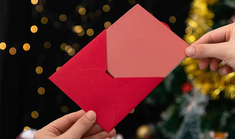 Two hands holding a red envelope against a festive background with lights and a Christmas tree.
