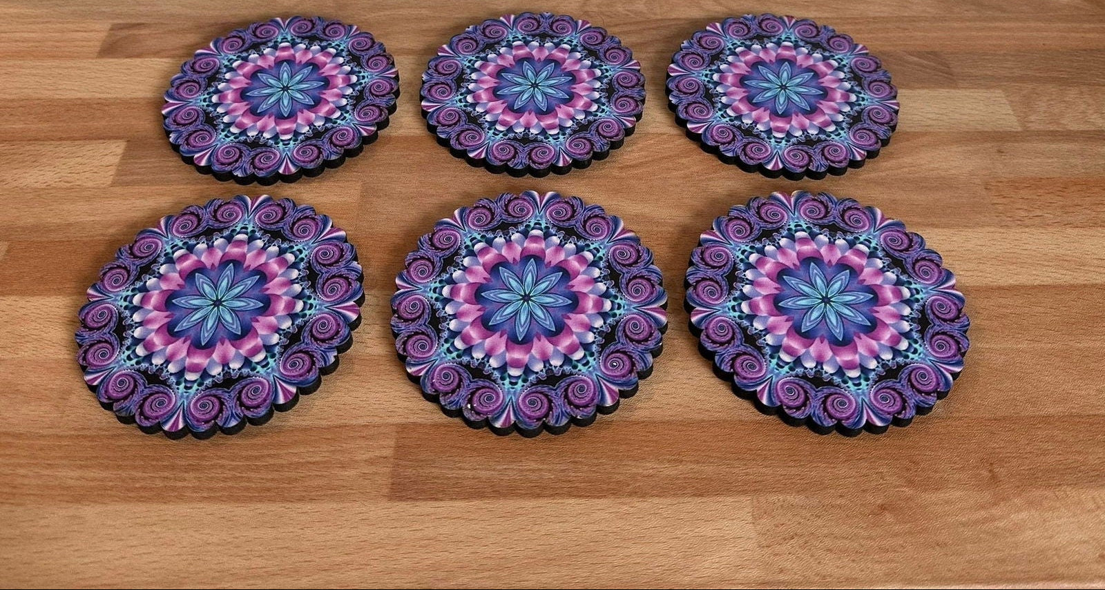 Set of six round purple and pink mandala coasters on a wooden surface with a geometric patterned rug in the background.