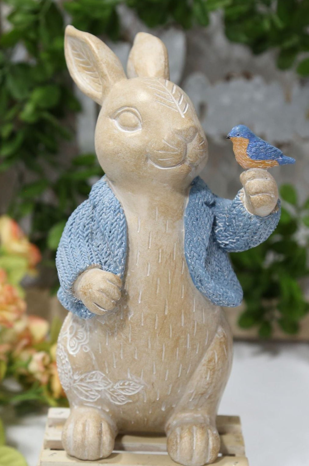 Statue of a rabbit in a blue outfit holding a flower, with a floral and wooden frame background.