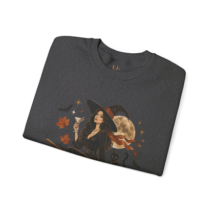 A dark heather gray folded t-shirt featuring a graphic print of a witch in a black hat and dress holding a glass, surrounded by autumn leaves, a full moon, stars, and a black cat, shown on a plain white background.