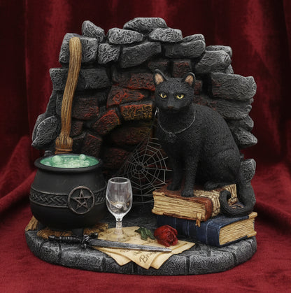 A gothic black cat fireplace figurine featuring a black cat sitting on old books in front of a stone fireplace with a cauldron, broom, wine glass, spiderweb, and a red rose.
