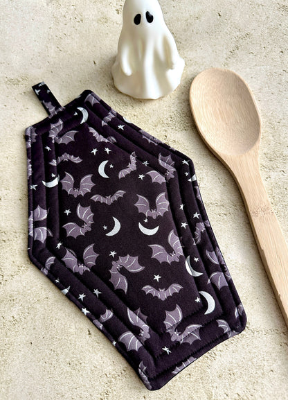 Pair of black pot holders with bat and moon pattern on a light surface with a wooden spoon and ghost decoration.
