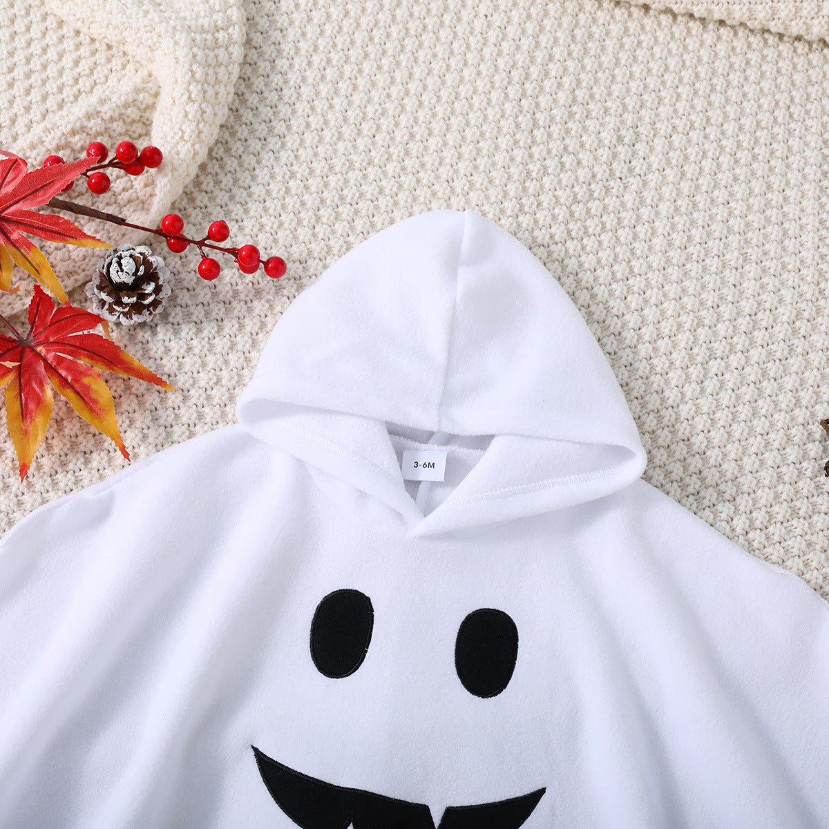 White hoodie with ghost face design on a textured surface with autumn leaves and pinecones.