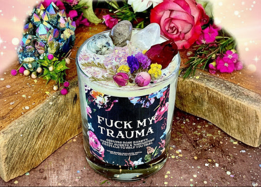 Candle with decorative elements and 'Fuck My Trauma' label on a wooden surface with flowers and crystals.