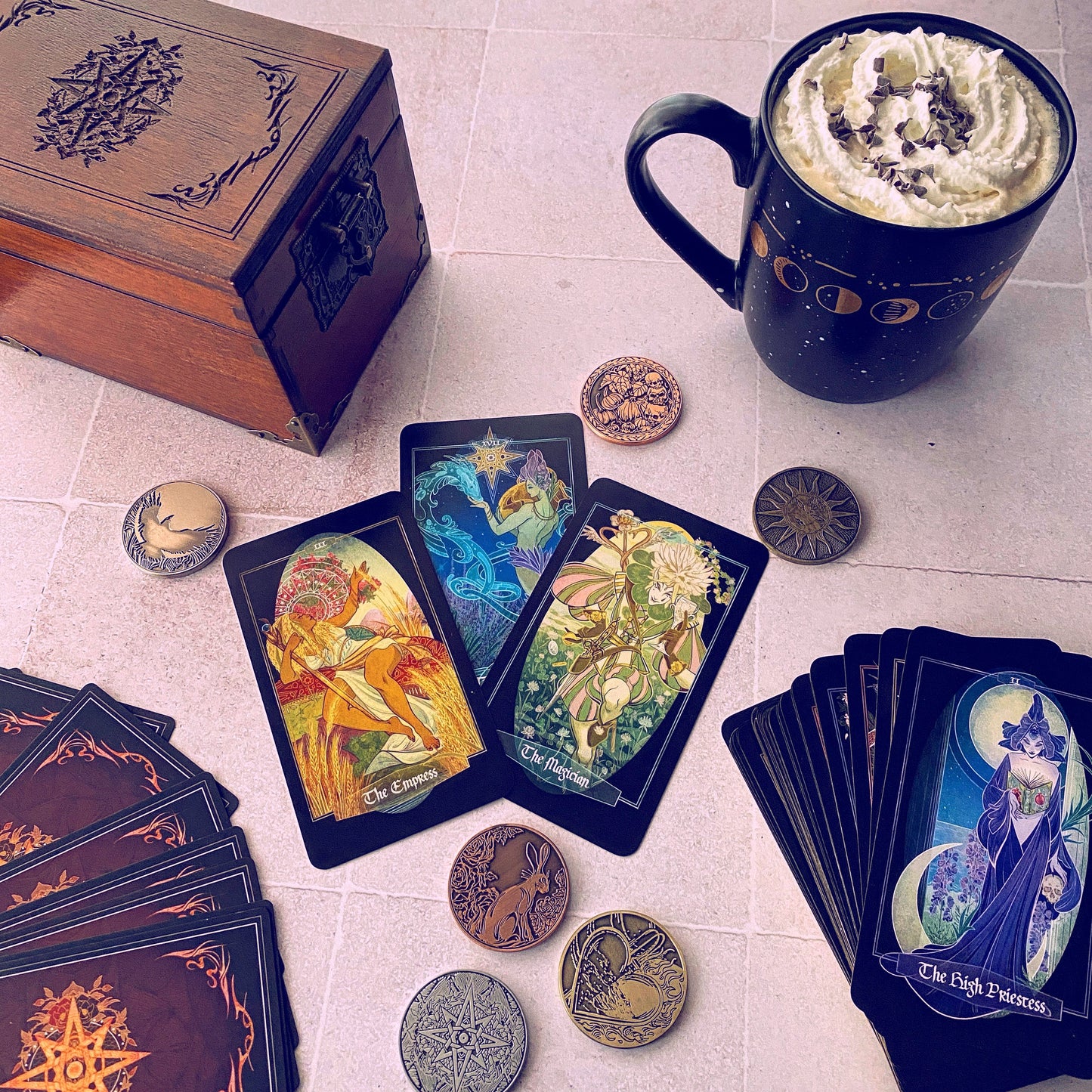 A styled flat-lay of tarot cards and themed accessories on a light tile surface. Visible items include multiple tarot card decks, several cards fanned out, several round metallic coin-like tokens, a dark wooden box with an engraved lid, and a black ceramic mug filled with whipped cream and chocolate drizzle.