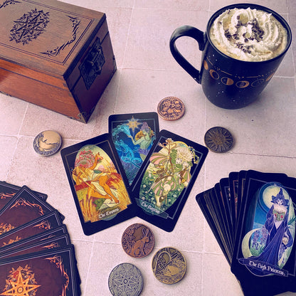 A styled flat-lay of tarot cards and themed accessories on a light tile surface. Visible items include multiple tarot card decks, several cards fanned out, several round metallic coin-like tokens, a dark wooden box with an engraved lid, and a black ceramic mug filled with whipped cream and chocolate drizzle.