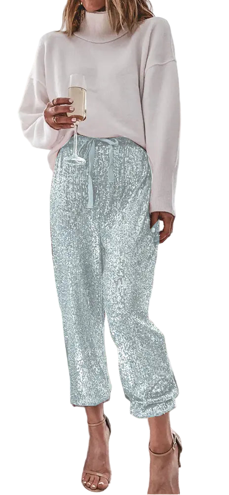 Woman in light blue sequin pants and white sweater holding a glass in a living room with a Christmas tree.