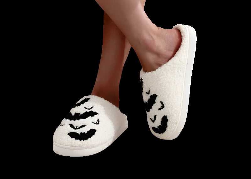 White slippers with black cat face design worn by a person sitting on a pink cushioned chair.