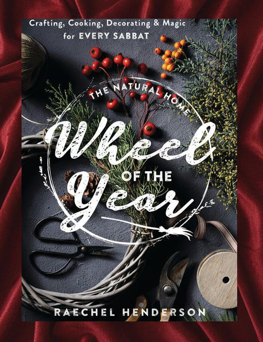 Book cover for 'The Natural Home: Wheel of the Year' by Raechel Henderson, featuring a dark slate background decorated with evergreen branches, red berries, pinecones, scissors, and a spool of thread.