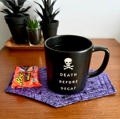 Black mug with 'Death Before Decaf' text on a purple coaster with a Halloween theme, next to a packet of Reese's Cups.