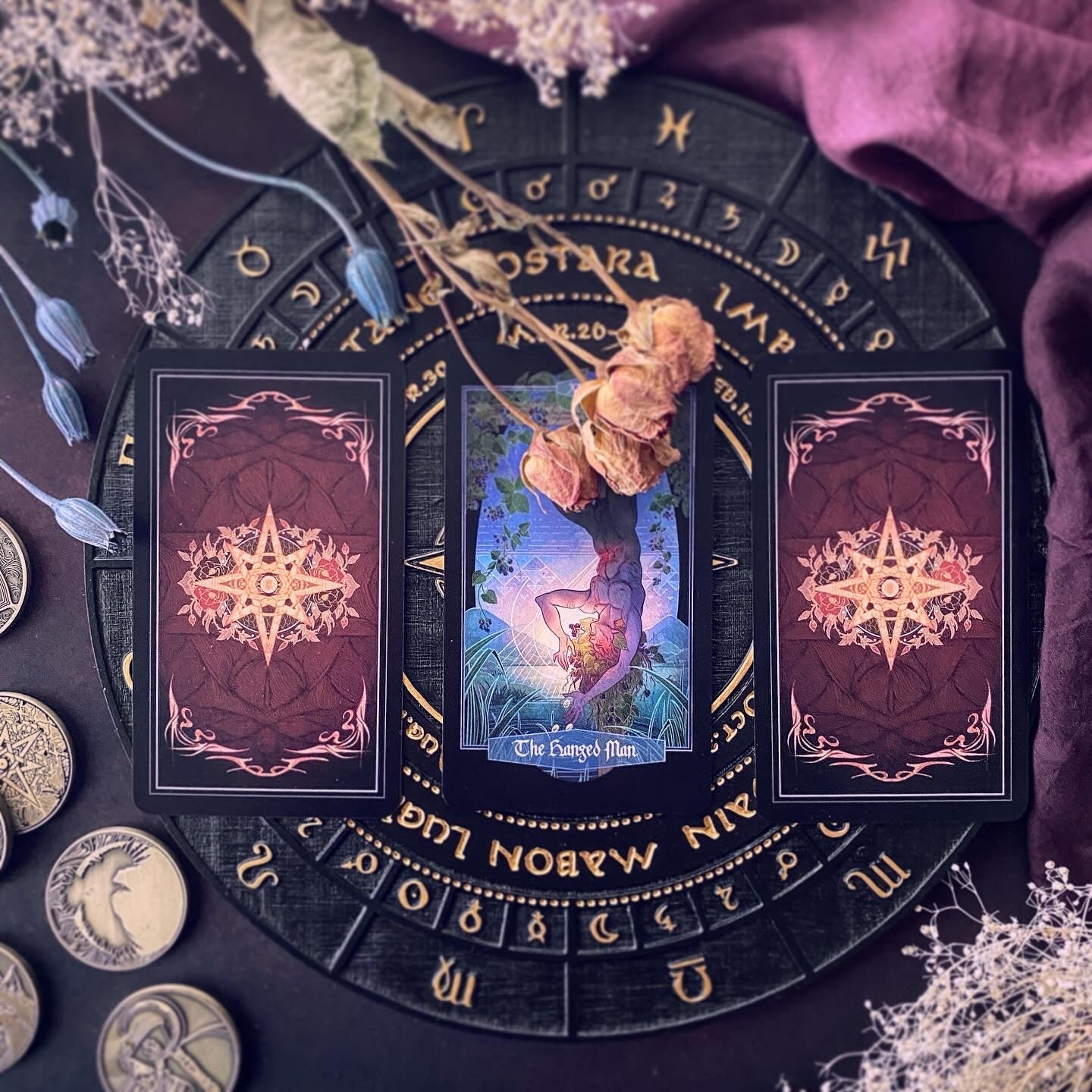 A tarot card deck arranged on a black circular astrological wheel with gold symbols, surrounded by dried flowers, coins, and purple fabric. The center card is 'The Chosen Path' showing a figure in a glowing blue and purple landscape, flanked by two back-facing cards with ornate gold star designs on a dark red background.