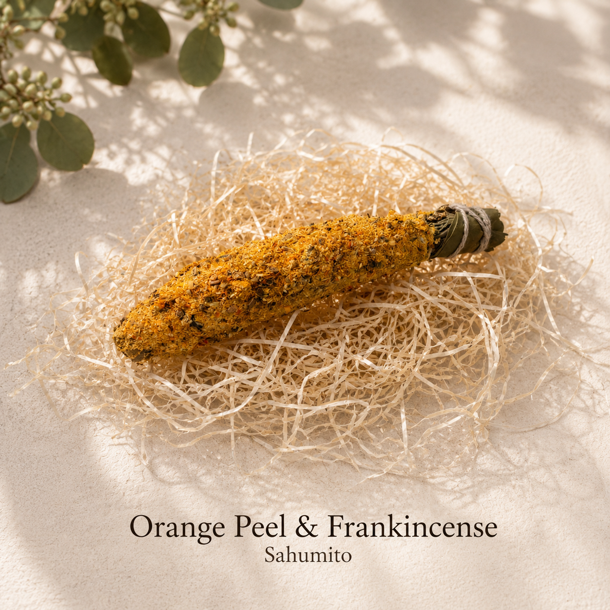 A dried orange peel and frankincense incense stick rests on a bed of shredded natural paper on a light beige surface, with soft leaf shadows and a few green leaves in the top corner. Text at the bottom reads 'Orange Peel & Frankincense' and 'Sahumito'.