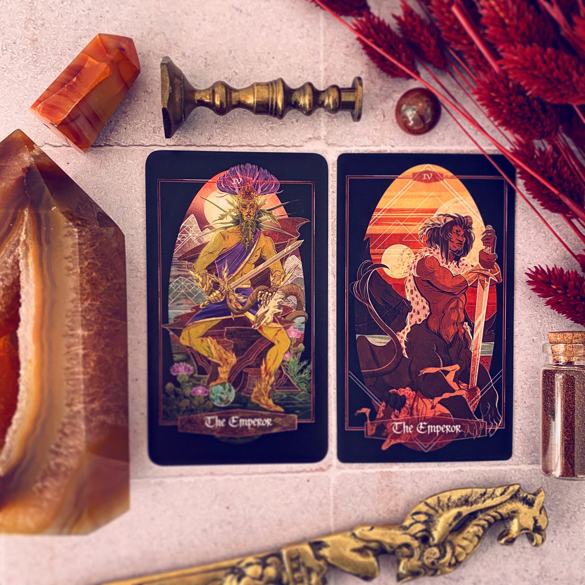 Two tarot cards titled 'The Emperor' are arranged on a light stone surface with decorative props including crystals, dried red foliage, a brass candlestick, and a small glass jar.