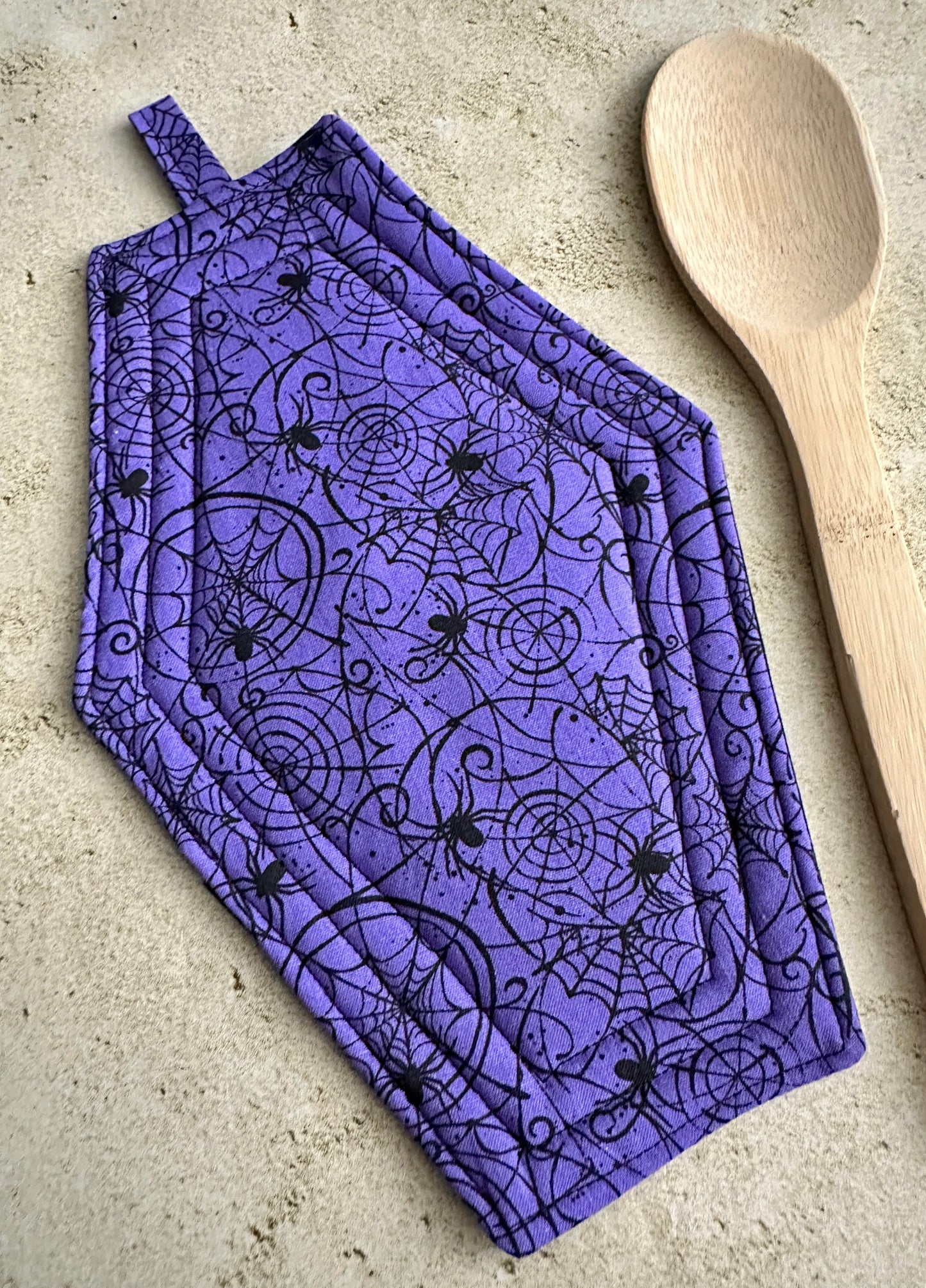 Purple pot holder with black spider design on a beige surface with a wooden spoon.
