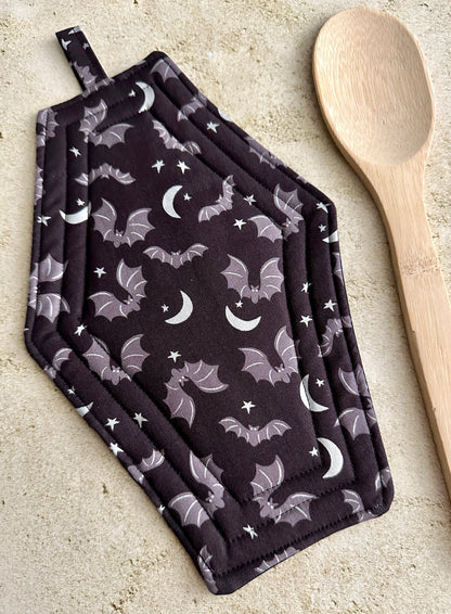 Set of black pot holders with bat and moon pattern on a light surface with a wooden spoon.
