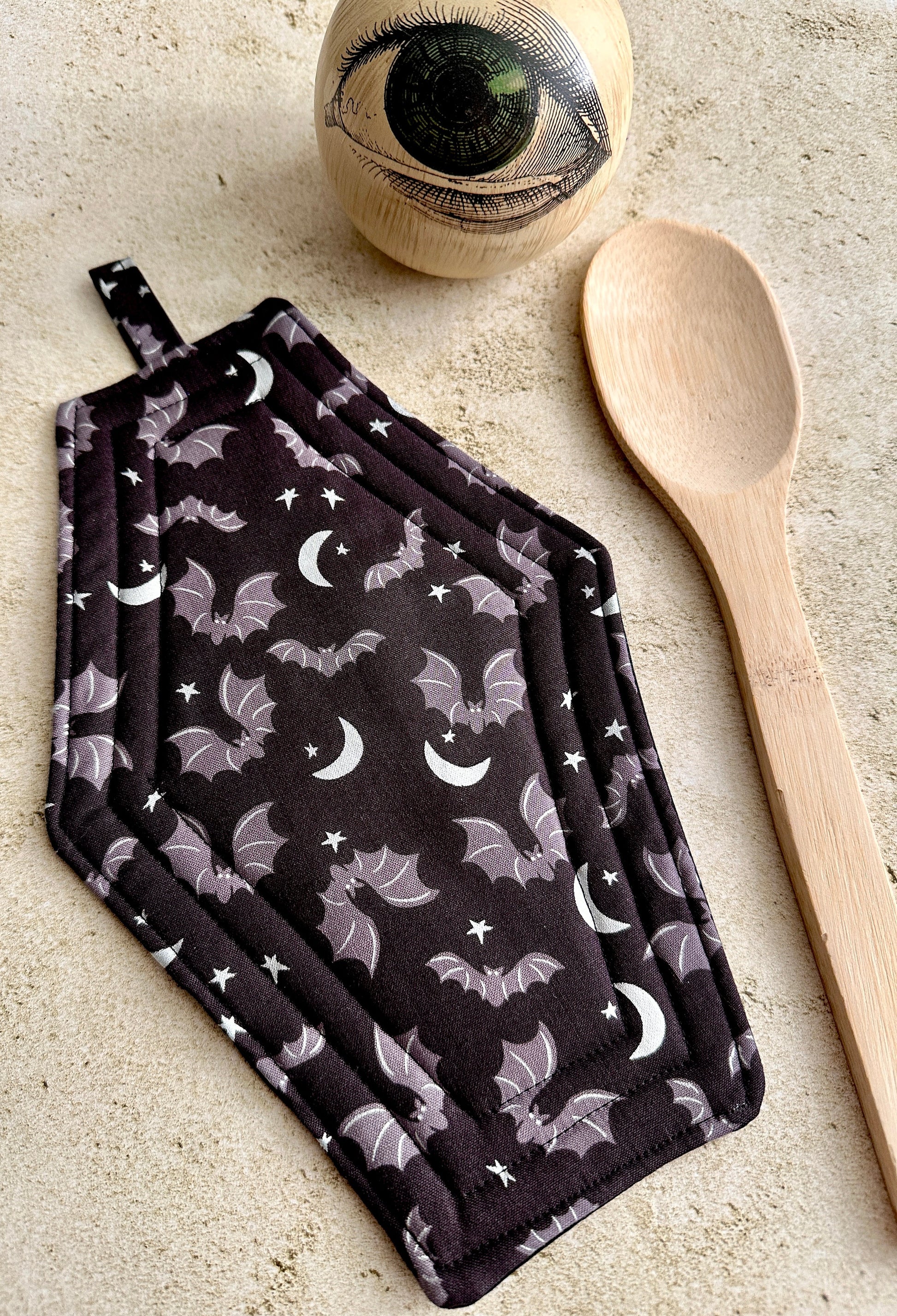 Pair of pot holders with bat and moon pattern on a neutral surface with a wooden spoon and eye-shaped object.