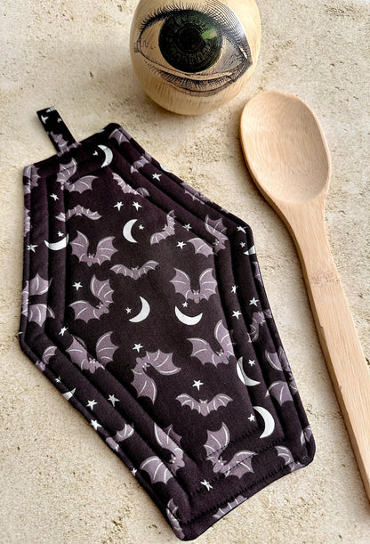 Pair of pot holders with bat and moon pattern on a neutral surface with a wooden spoon and eye-shaped object.