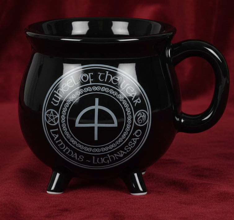 A glossy black ceramic mug shaped like a cauldron with three feet, featuring a white printed 'Wheel of the Year' symbol and text on the front, set against a plain white background.