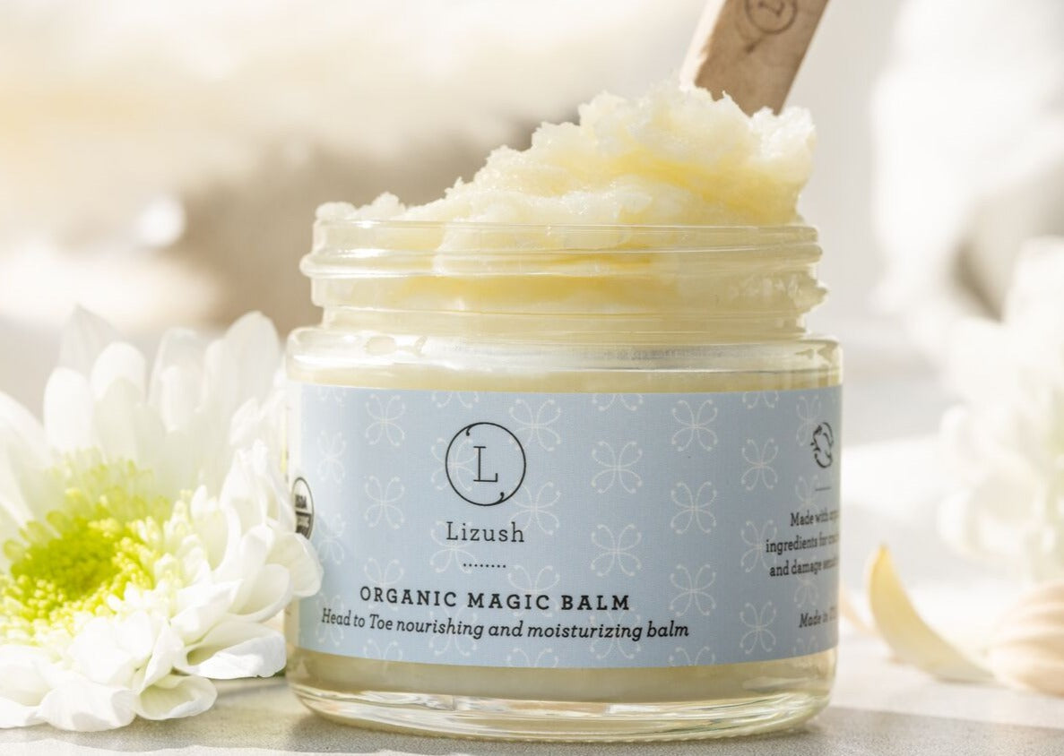 Jar of Lizush Organic Magic Balm with a wooden applicator against a blurred floral background