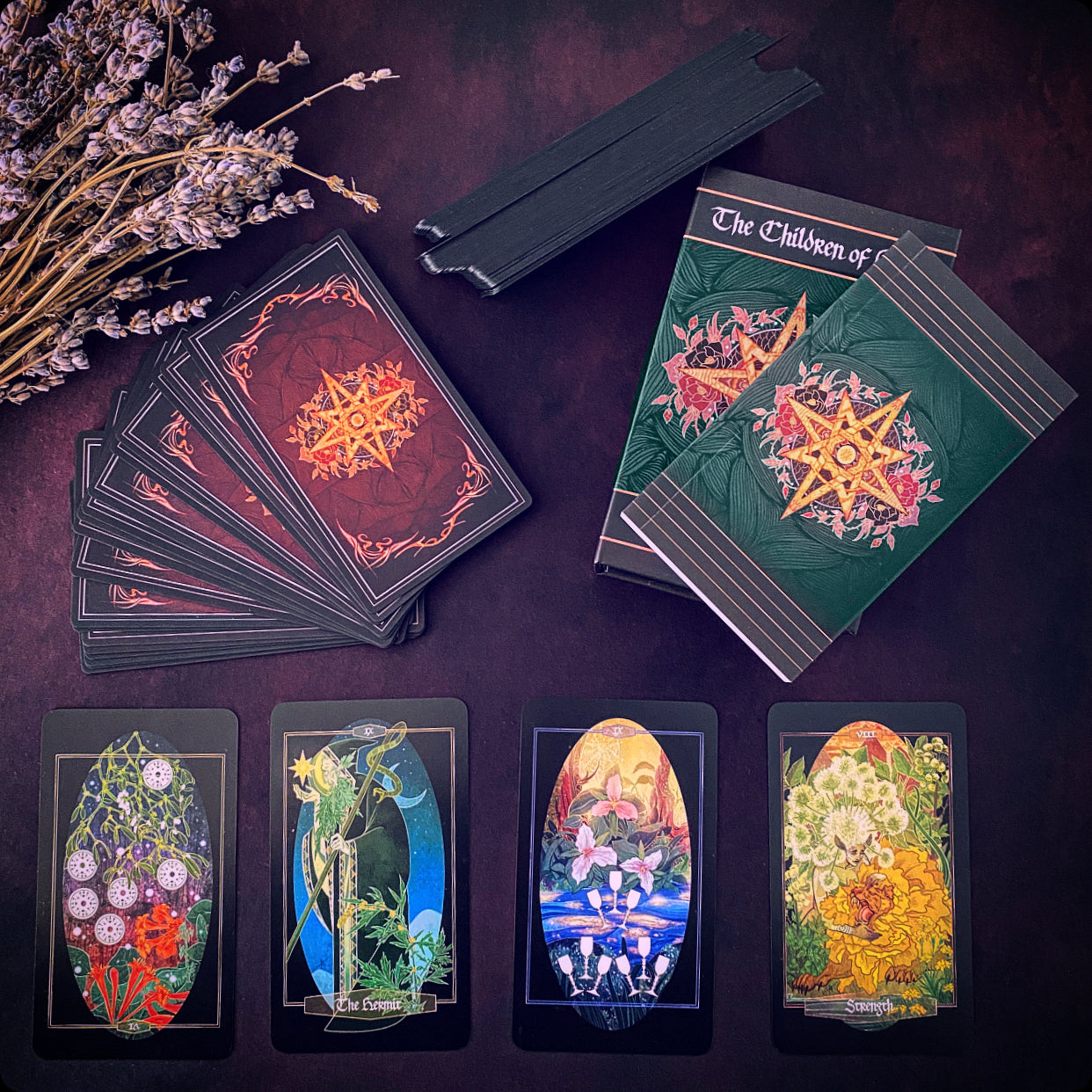 A tarot card deck set arranged on a dark surface, including a closed box, a fanned deck of cards, a stack of cards, and four individual cards. The packaging and card backs feature ornate red and green designs with gold star-like symbols, while the face-up cards display colorful fantasy-style illustrations.