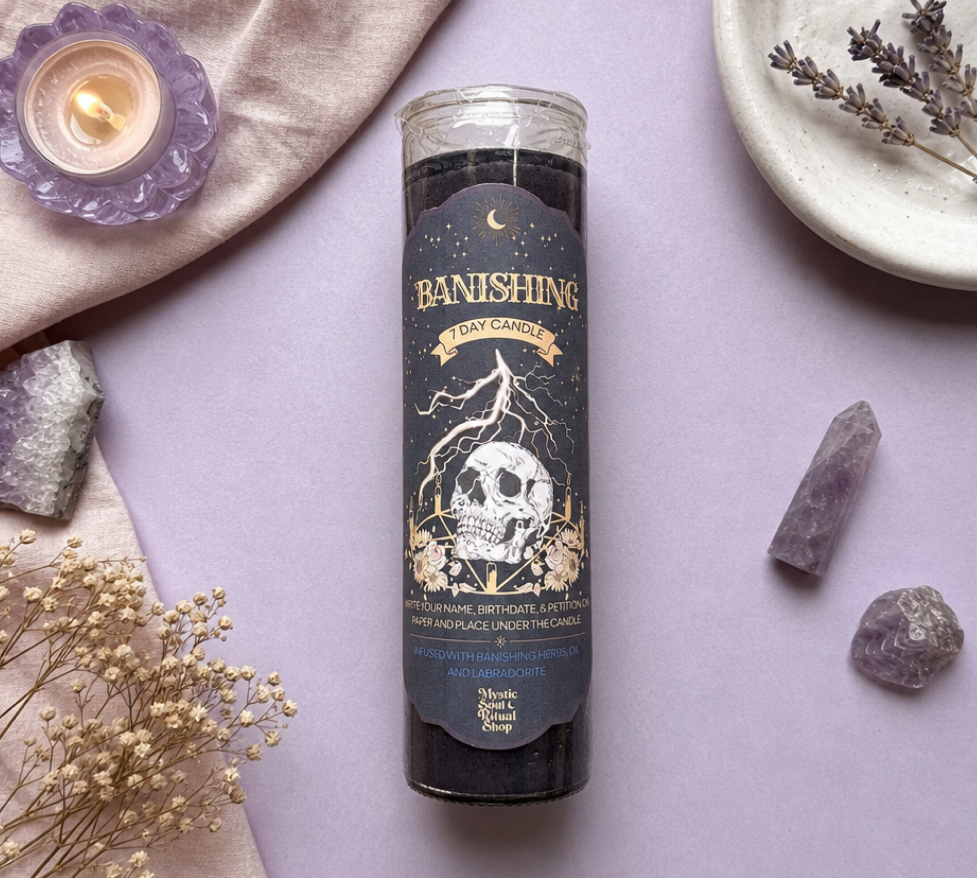 A black tarot card candle in a clear glass jar labeled 'Banishing Day Candle' by Magic Shop, styled in a flat lay on a lavender background with crystals, dried flowers, and a small lit candle.
