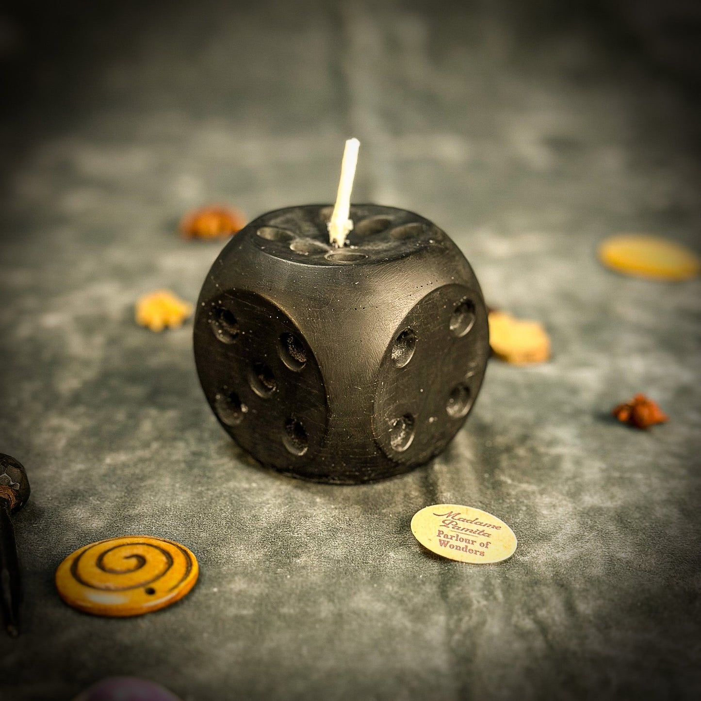 Black dice-shaped candle with a lit wick on a dark surface with decorative items.