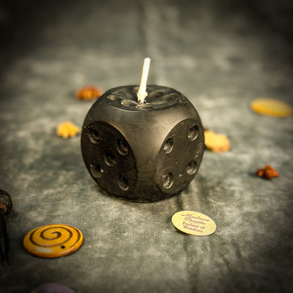 Black dice-shaped candle with a lit wick on a dark surface with decorative items.