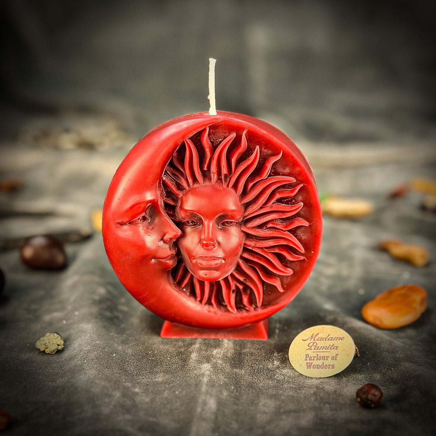 Red candle with sun and moon design on a dark surface, featuring a brand label.