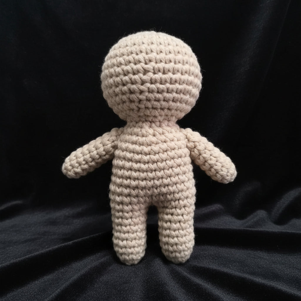 A handmade beige crochet doll with a round head, short arms, and legs, standing on a gray surface in front of a window.