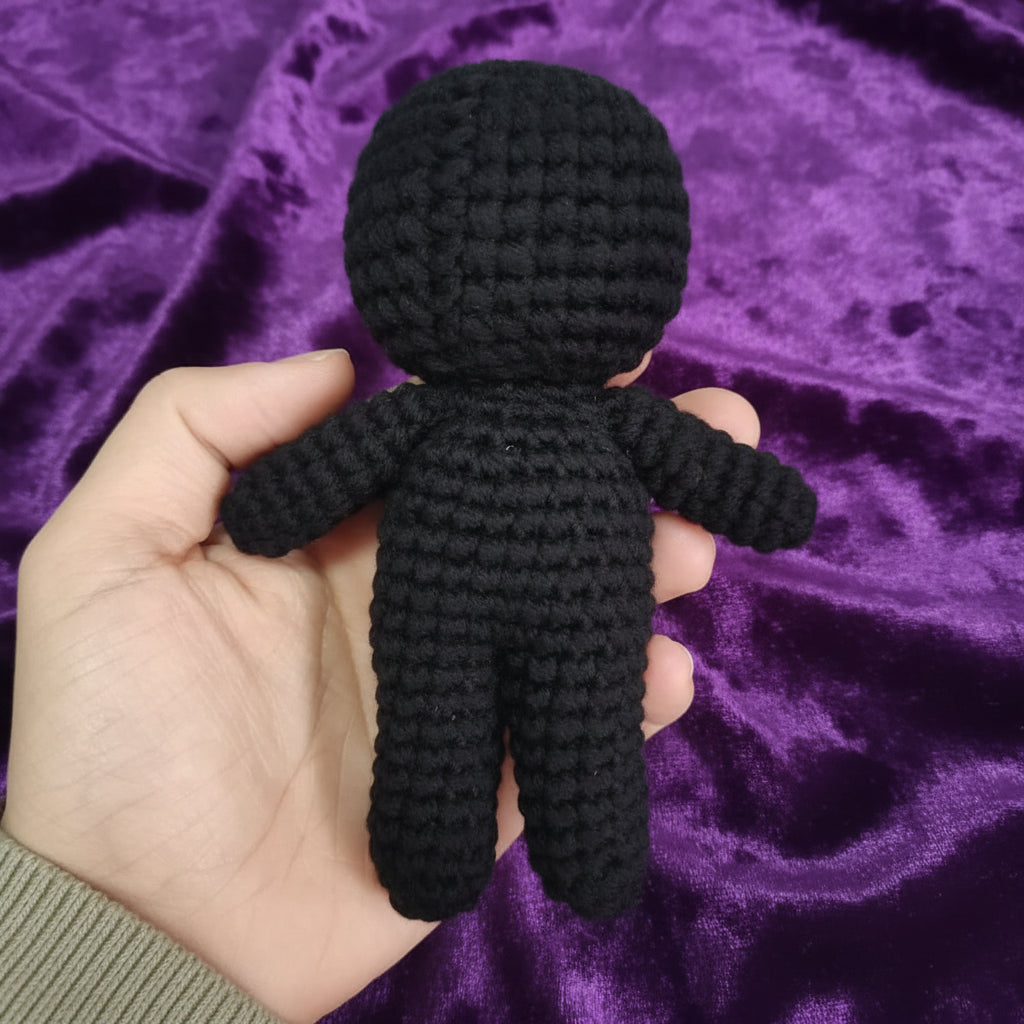 A hand holding a small black crocheted doll with a round head, simple arms, and legs against a light gray background.