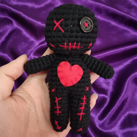 Black crochet doll with red heart and stitches held in a hand against a light background