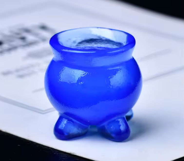 A small glossy cobalt blue glass cup with a rounded body and three short feet, photographed on a white paper surface.