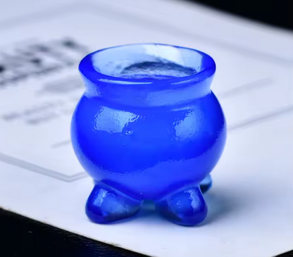 A small glossy cobalt blue glass cup with a rounded body and three short feet, photographed on a white paper surface.