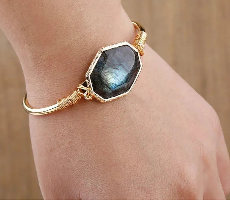 A gold bangle bracelet with a large hexagonal dark gray labradorite stone, worn on a person's wrist against a warm wooden background.