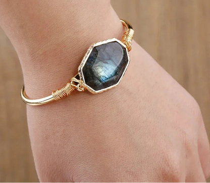 A gold bangle bracelet with a large hexagonal dark gray labradorite stone, worn on a person's wrist against a warm wooden background.