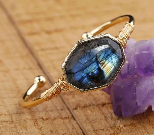 A gold-toned open bangle ring featuring a large hexagonal labradorite stone with blue, black, and gold iridescence, styled on a wooden surface next to a purple crystal cluster.