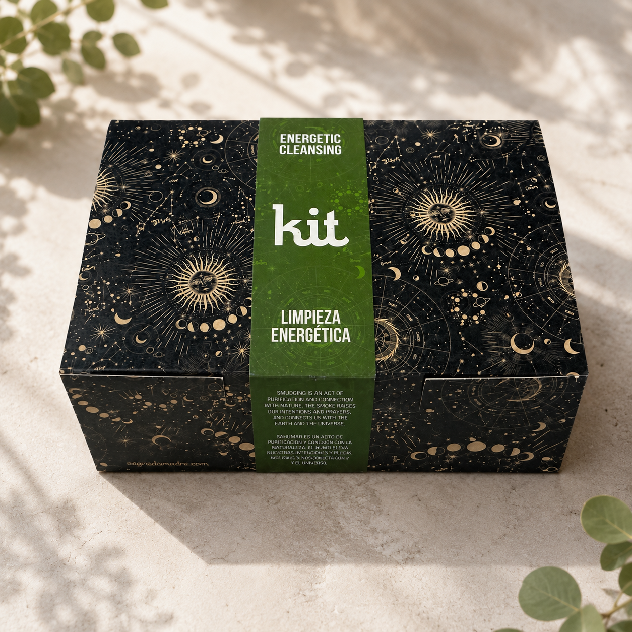 A rectangular black box with a green vertical band labeled 'kit' and 'ENERGETIC CLEANSING / LIMPIEZA ENERGETICA' sits on a light stone surface with soft leaf shadows and greenery around it. The box lid is decorated with gold celestial patterns including suns, moons, and stars.
