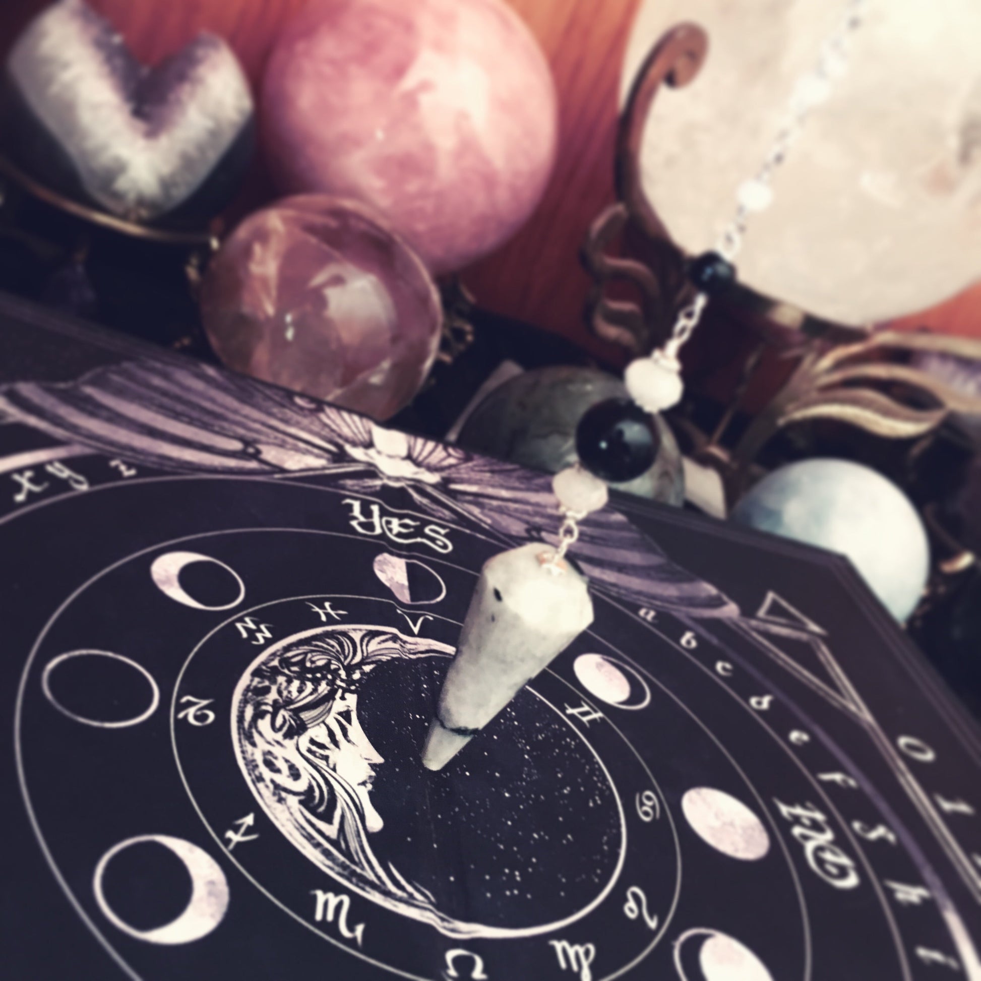 A close-up of a celestial-themed pendant necklace featuring a white crystal cone and black beads on a silver chain, resting on a black astrology chart surrounded by crystals and mystical props.