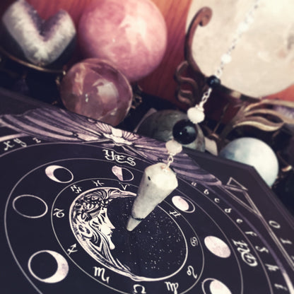 A close-up of a celestial-themed pendant necklace featuring a white crystal cone and black beads on a silver chain, resting on a black astrology chart surrounded by crystals and mystical props.