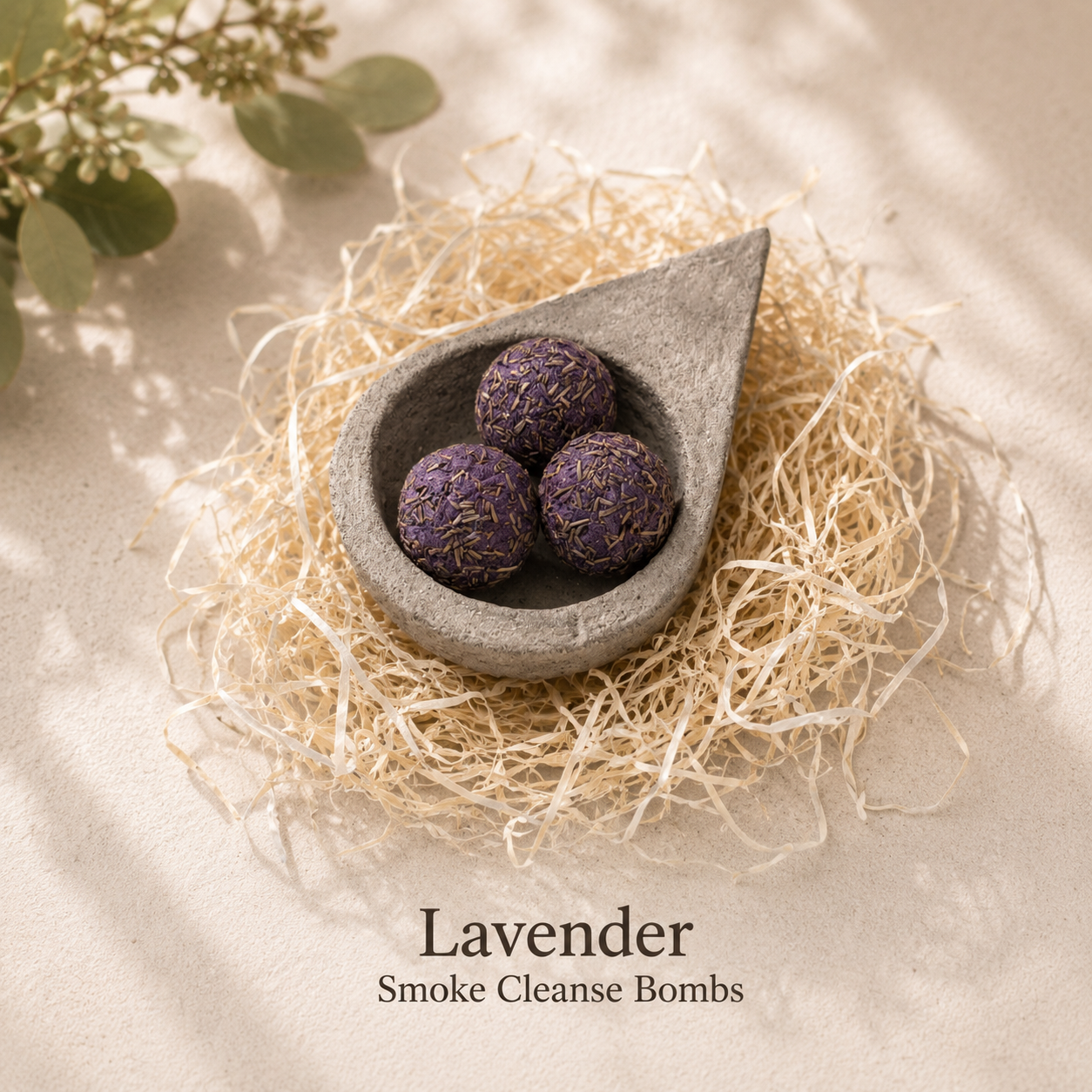 Three lavender smoke cleanse bombs in a dark gray teardrop-shaped bowl, set on a bed of natural raffia fiber. The scene is styled with soft-focus eucalyptus leaves in the top left corner and text at the bottom reading 'Lavender Smoke Cleanse Bombs'.