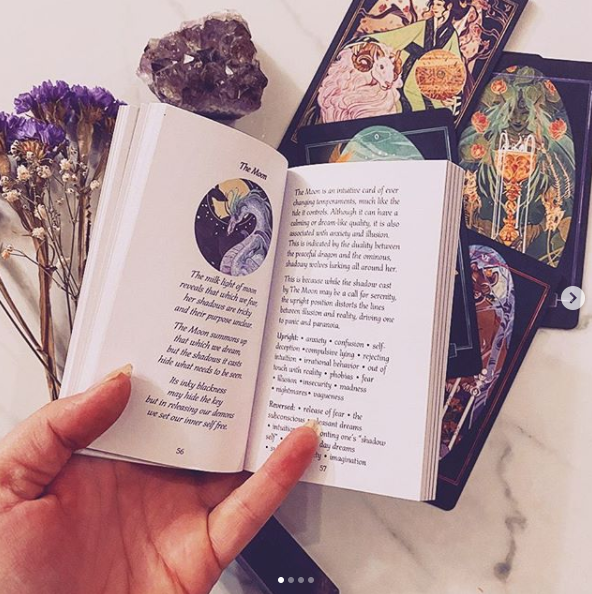 An open book with a poem titled 'The Moon' on one page, held by a hand, surrounded by tarot cards and dried purple flowers on a light marble surface.