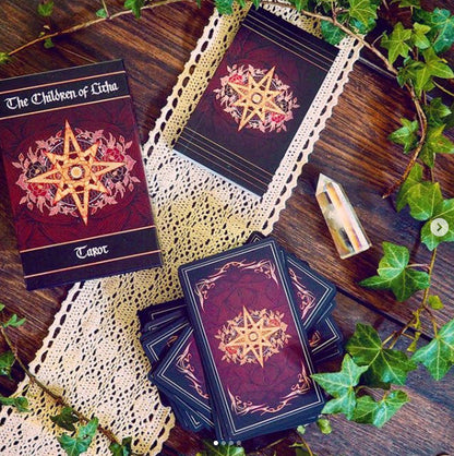 A tarot deck set arranged on a rustic wooden surface with cream lace doilies and green ivy vines. The box and cards show a dark red and black background with a gold star design and floral accents.