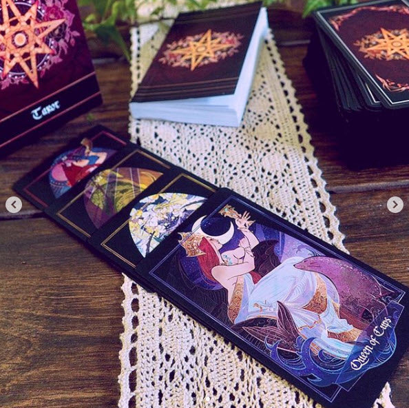 A tarot deck with illustrated cards arranged on a wooden table over a white lace runner. The front card reads "Queen of Cups," and other cards are partially visible, with the deck box and additional cards in the background.