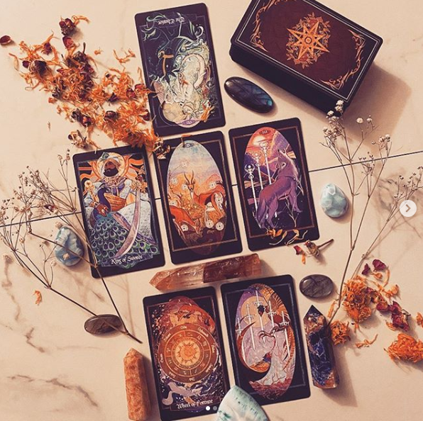 A set of illustrated tarot cards arranged on a light marble surface with dried flowers, crystals, and stones. The cards feature dark backgrounds with colorful mystical and celestial artwork, and a matching dark box with an ornate gold emblem is visible at the top right.