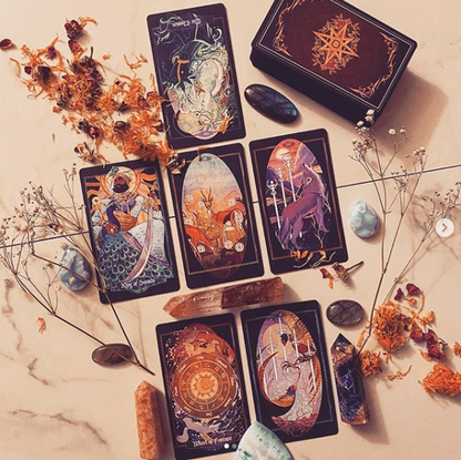 A set of illustrated tarot cards arranged on a light marble surface with dried flowers, crystals, and stones. The cards feature dark backgrounds with colorful mystical and celestial artwork, and a matching dark box with an ornate gold emblem is visible at the top right.