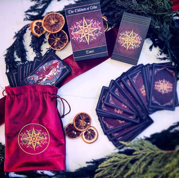 A tarot card deck set with a red drawstring bag, several cards fanned out, one upright card, and the box cover, arranged on a white surface with dried orange slices and evergreen branches.