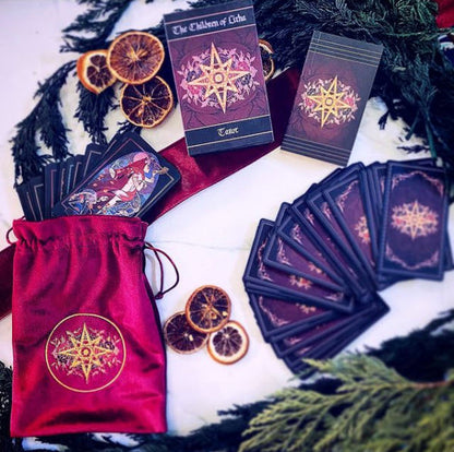 A tarot card deck set with a red drawstring bag, several cards fanned out, one upright card, and the box cover, arranged on a white surface with dried orange slices and evergreen branches.