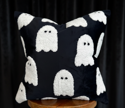 A square black throw pillow with a white textured ghost pattern, displayed on a wooden stool against a plain white background.