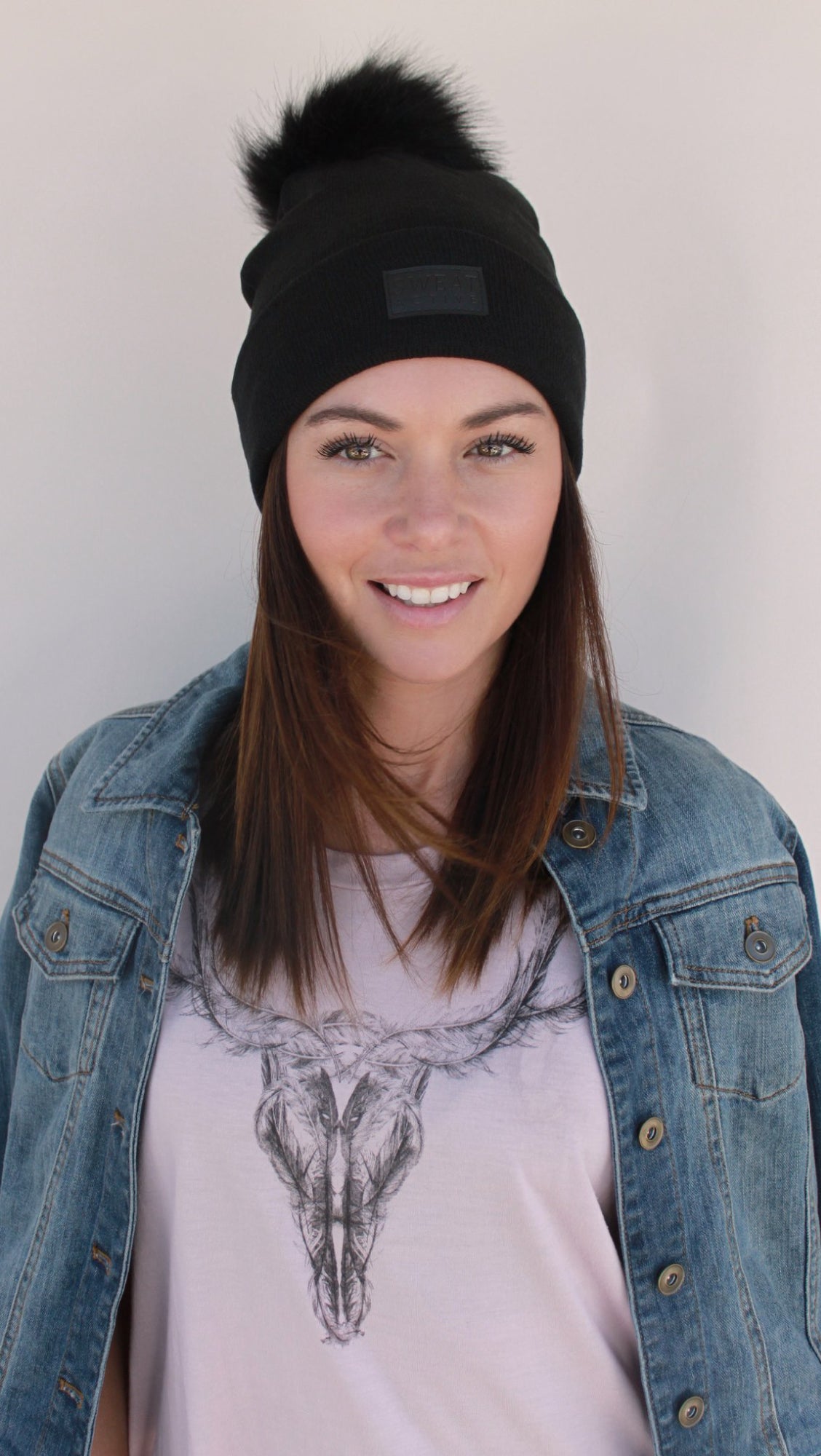 Woman wearing a black beanie with a pom-pom and denim jacket over a graphic t-shirt.