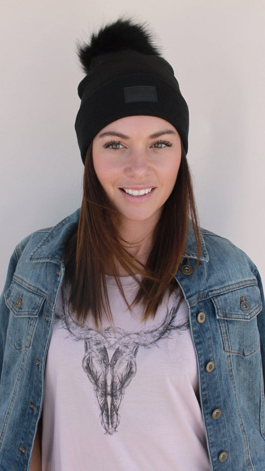 Woman wearing a black beanie with a pom-pom and denim jacket over a graphic t-shirt.